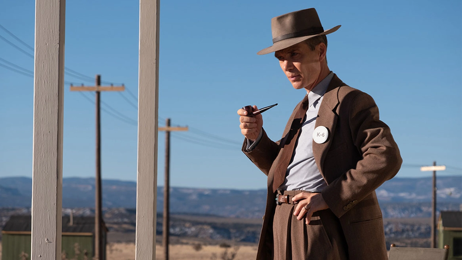 Actor Cillian Murphy as J. Robert Oppenheimer in the 2023 film, dressed in a brown suit and hat, holding a pipe and standing in an outdoor setting with a row of power pylons to the left of the image